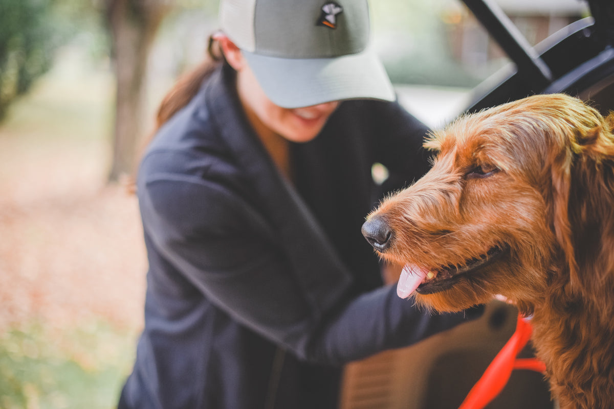 A woman wearing a cap smiles as she stands next to a happy brown dog sitting in the open trunk of a car, with outdoor gear packed inside and green trees and fallen leaves in the background.