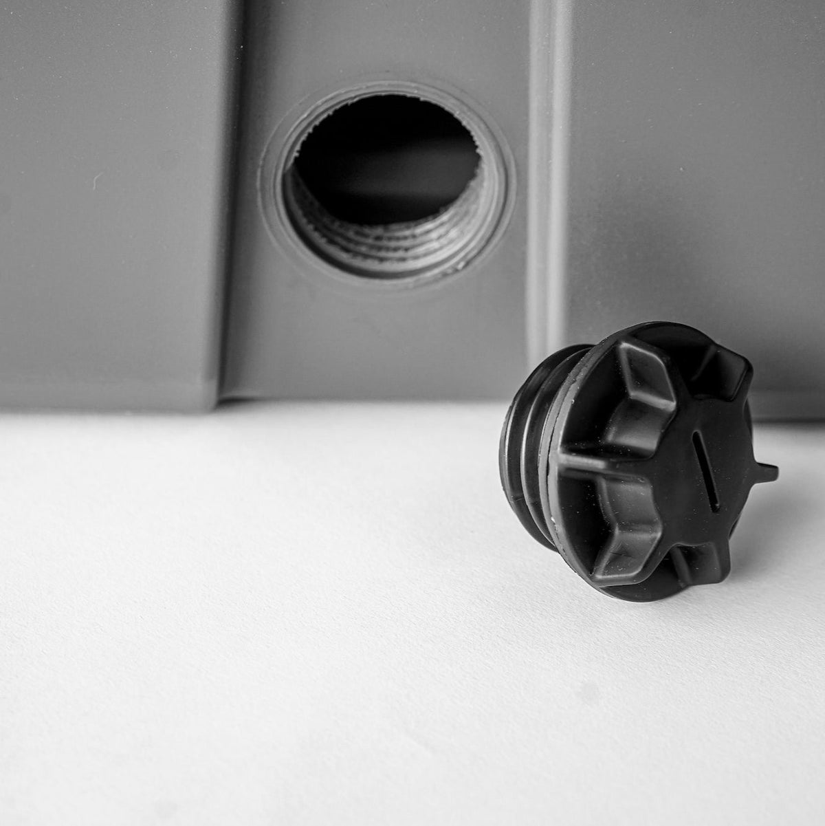 A close-up of the black GUNNER DRAIN PLUG 2.0 screw cap beside its threaded opening on a gray GUNNER kennel, shown on a white surface.