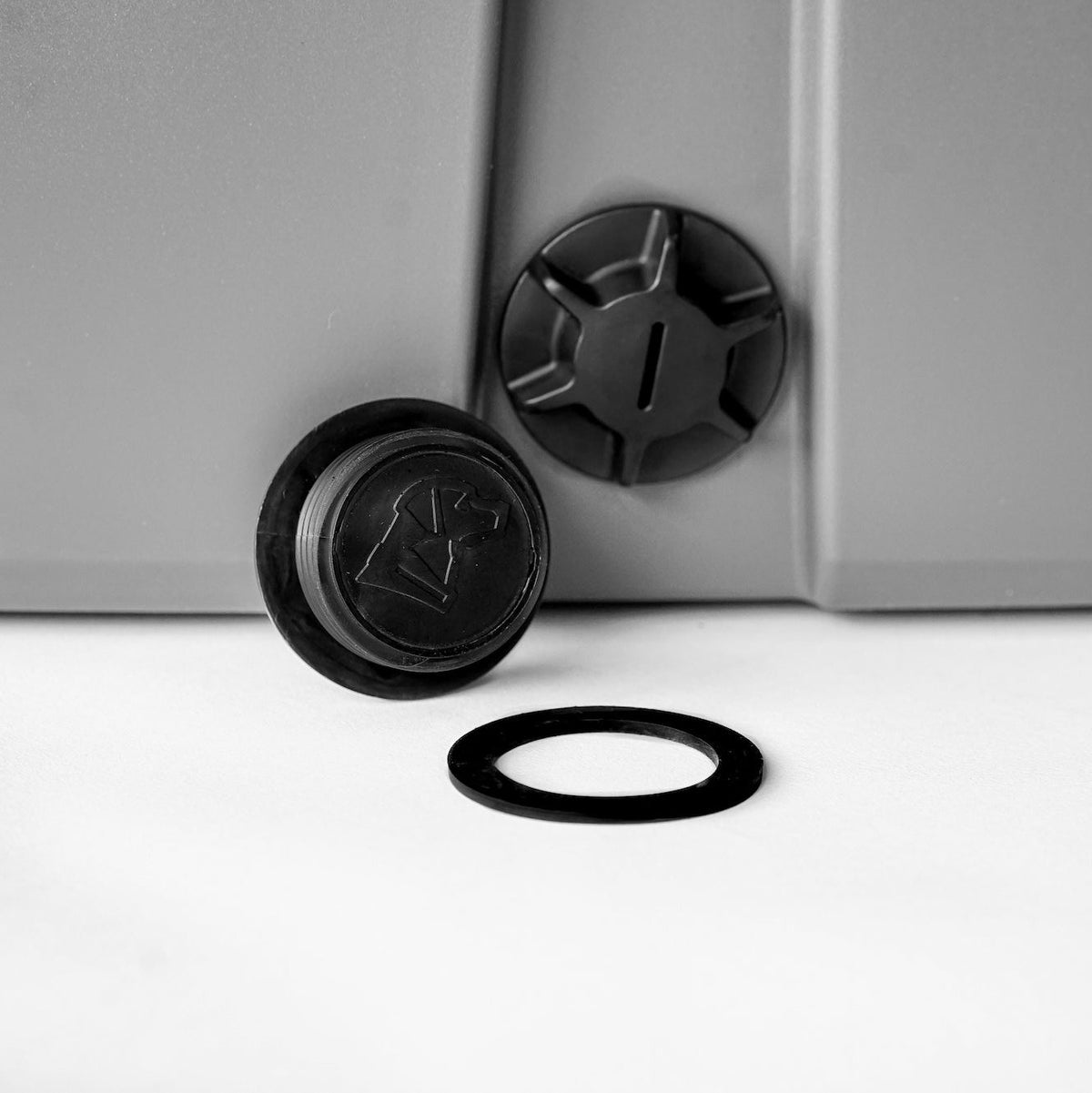 A close-up of two black GUNNER DRAIN PLUG 2.0 plastic plugs and a rubber washer on a gray surface, with one plug detached next to the washer on a white background.