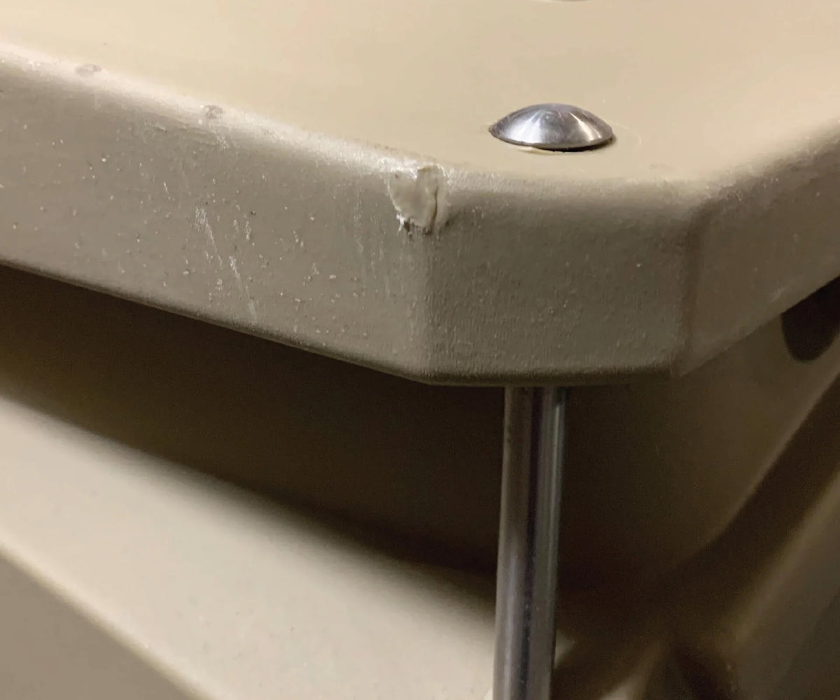 A close-up of a beige plastic dog crate surface with a small chipped area on the corner, showing minor damage. A metal screw and a metal rod are also visible near the edge.