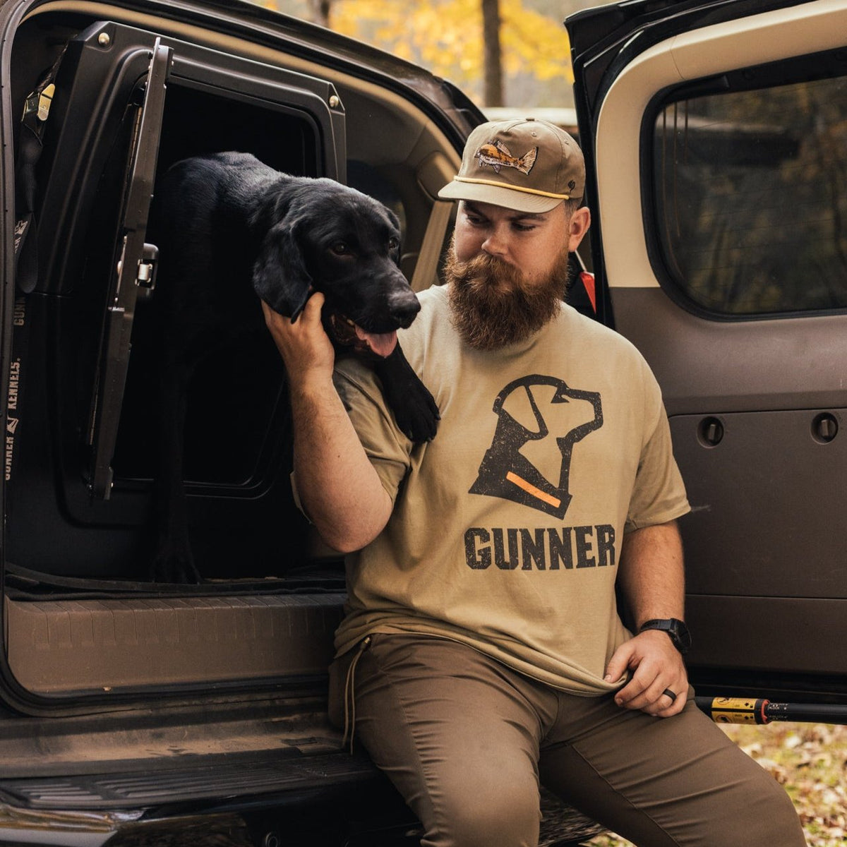 A bearded man in a tan GUNNER OG TEE SHIRT and cap sits on the back of an open vehicle, gently holding a black dog stepping out of a kennel, with autumn trees visible in the background.