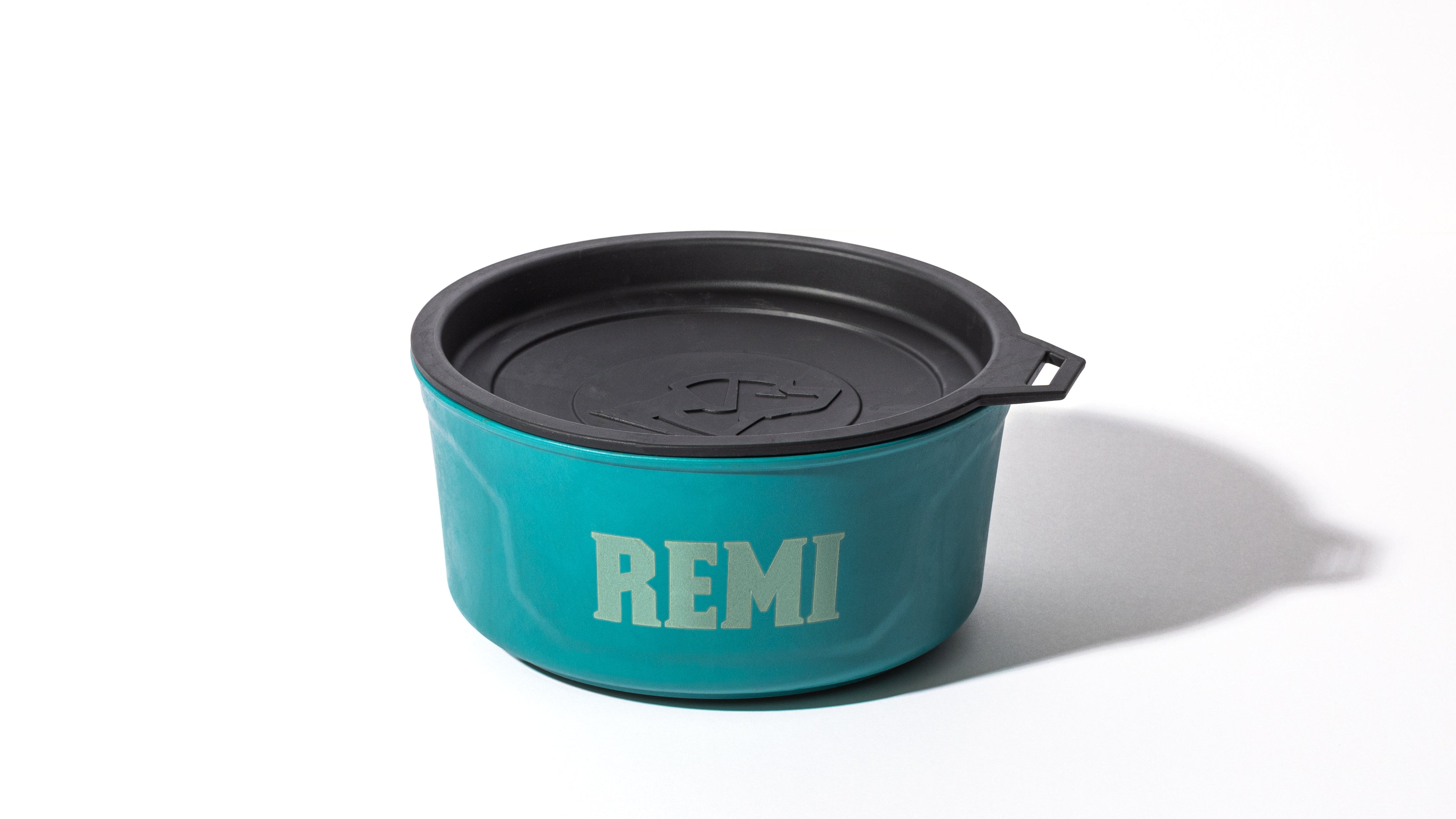 A teal container with a black lid featuring a handle and the word REMI printed in bold white letters on the side, perfect for use in a dog kennel or while traveling with your working dog, placed on a white surface.