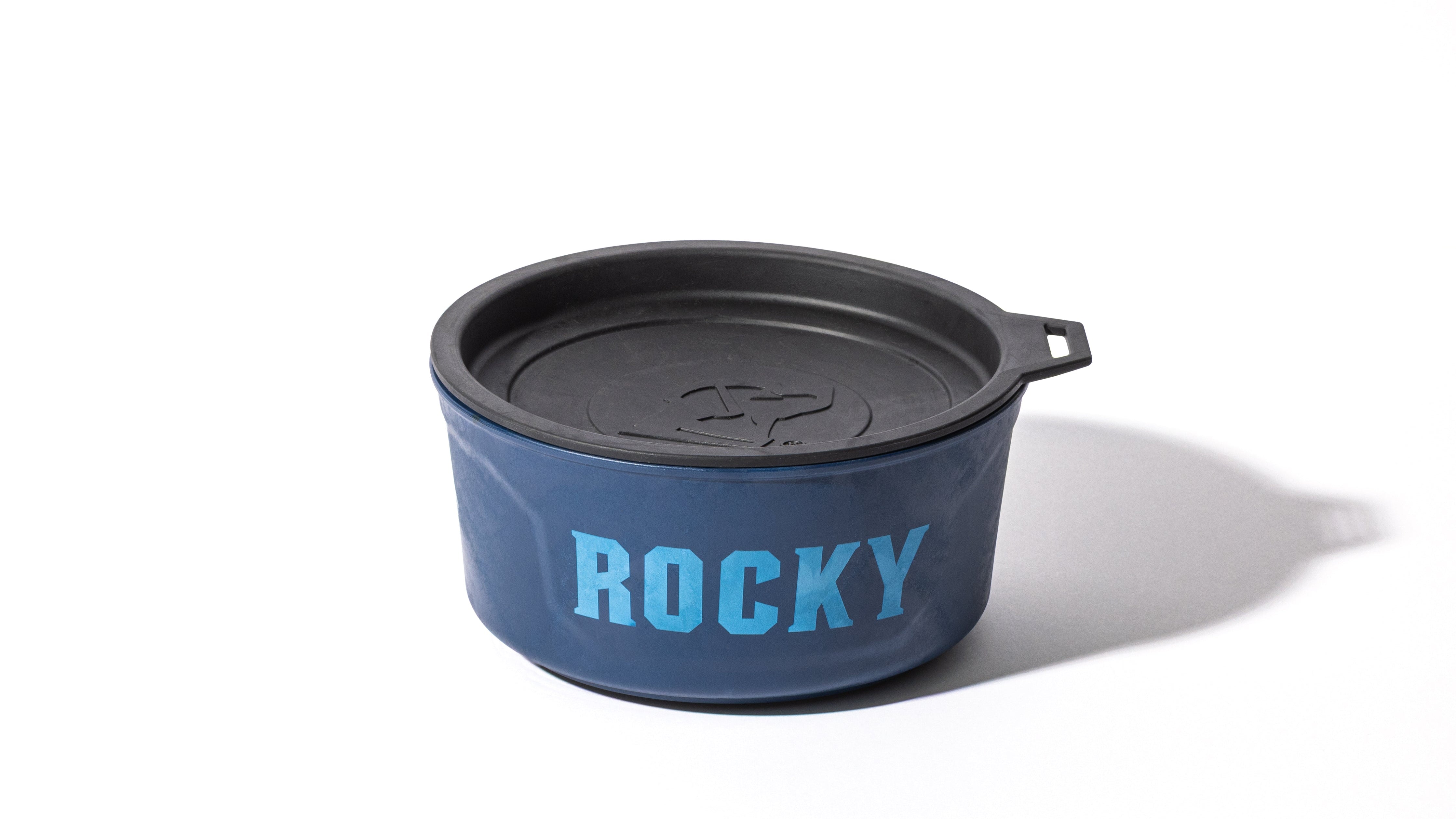 A blue food bowl with a black lid and handle, featuring the name ROCKY in large light blue letters on the front. Perfect for your hunting dog, this bowl is essential outdoor gear and casts a shadow on a white background.