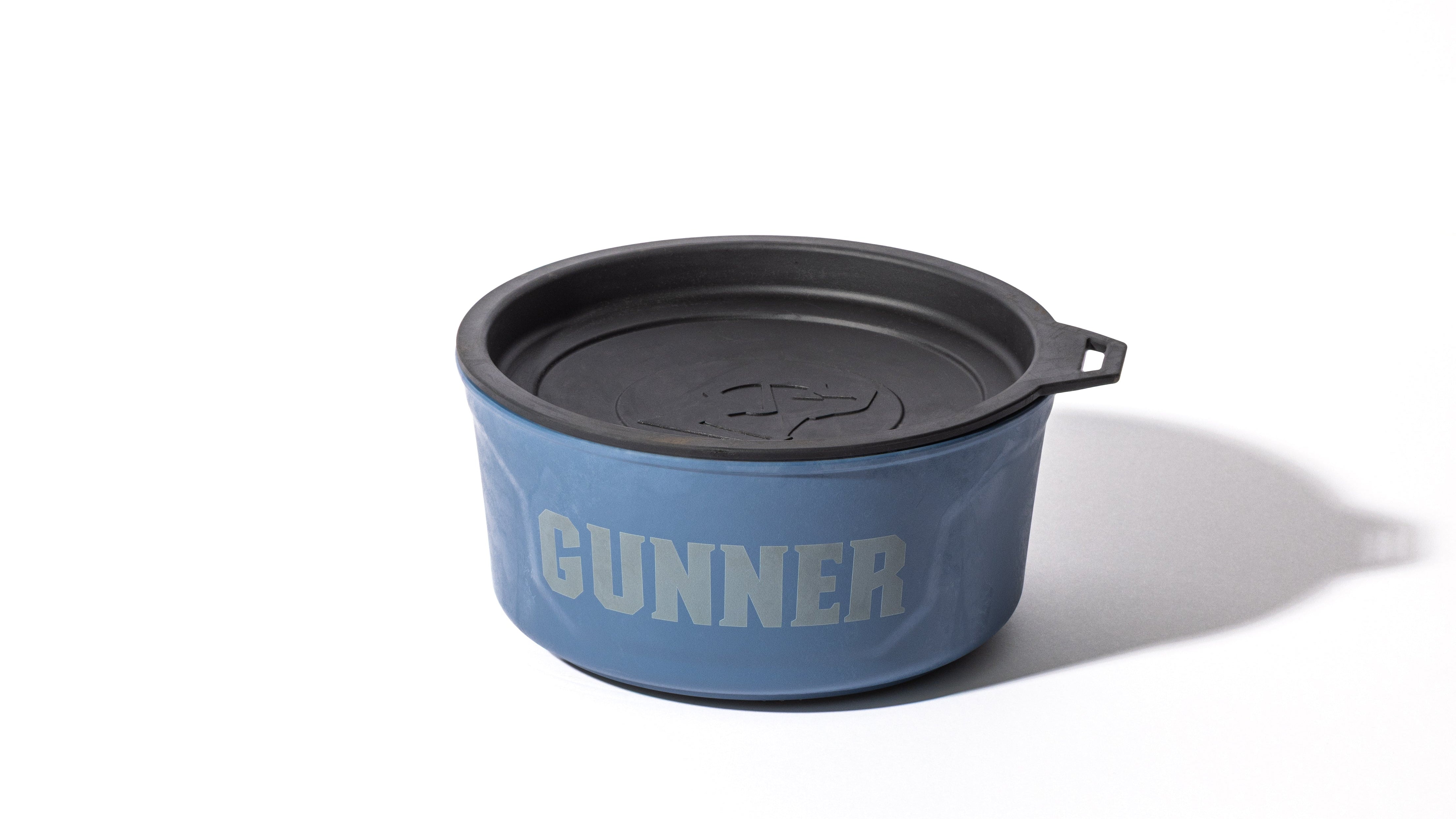 A blue pet bowl with a black lid, featuring the word GUNNER in large, bold letters on the side—perfect for pairing with your favorite dog crate or outdoor gear—placed on a white surface with a shadow to the right.