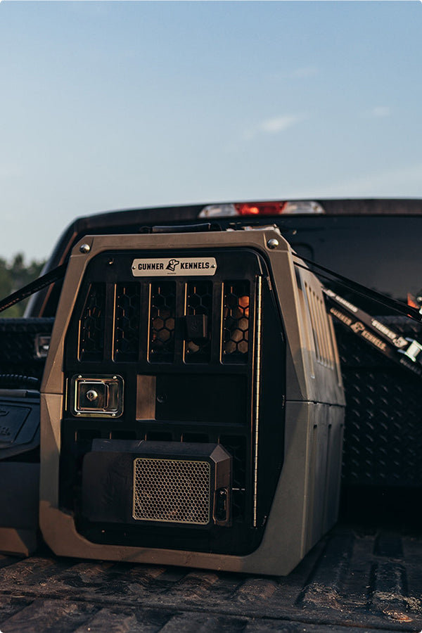 A sturdy Gunner Kennels hunting dog crate is secured in the bed of a pickup truck, ready for transport outdoors under a clear sky.