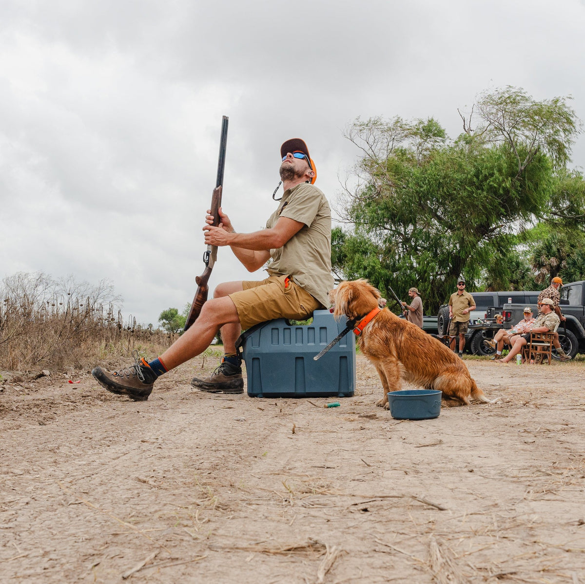 A man with ear protection and sunglasses sits on a GUNNER FOOD CRATE - Ducks Unlimited Blue by GUNNER, holding a shotgun. A brown dog is beside him on a dirt path, while people and vehicles are seen in the cloudy background.