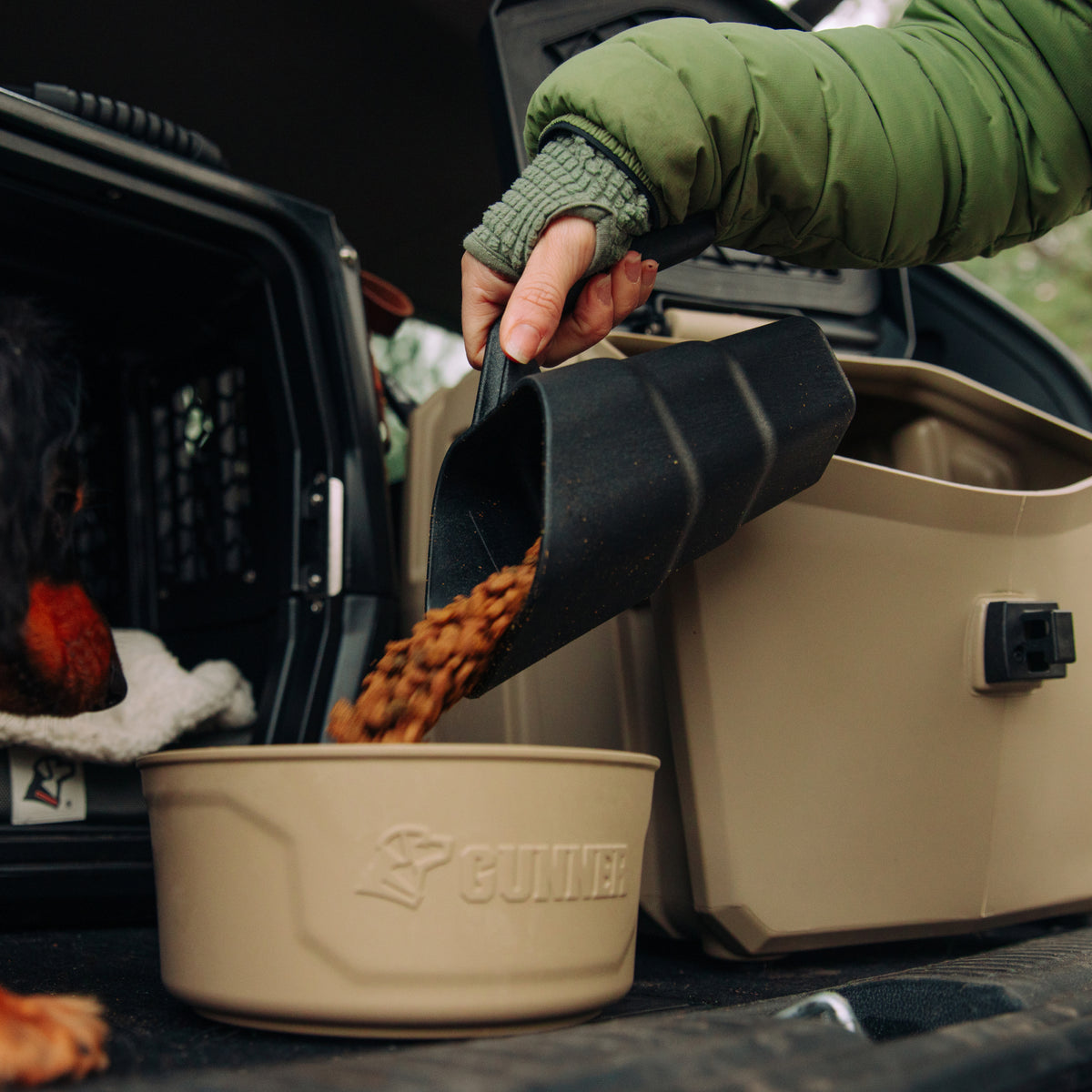 A person pours dry dog food from a black scoop into a beige GUNNER bowl from the Food & Water Bundle, with a dog in a crate nearby and storage containers behind them, highlighting GUNNERs organized feeding system.