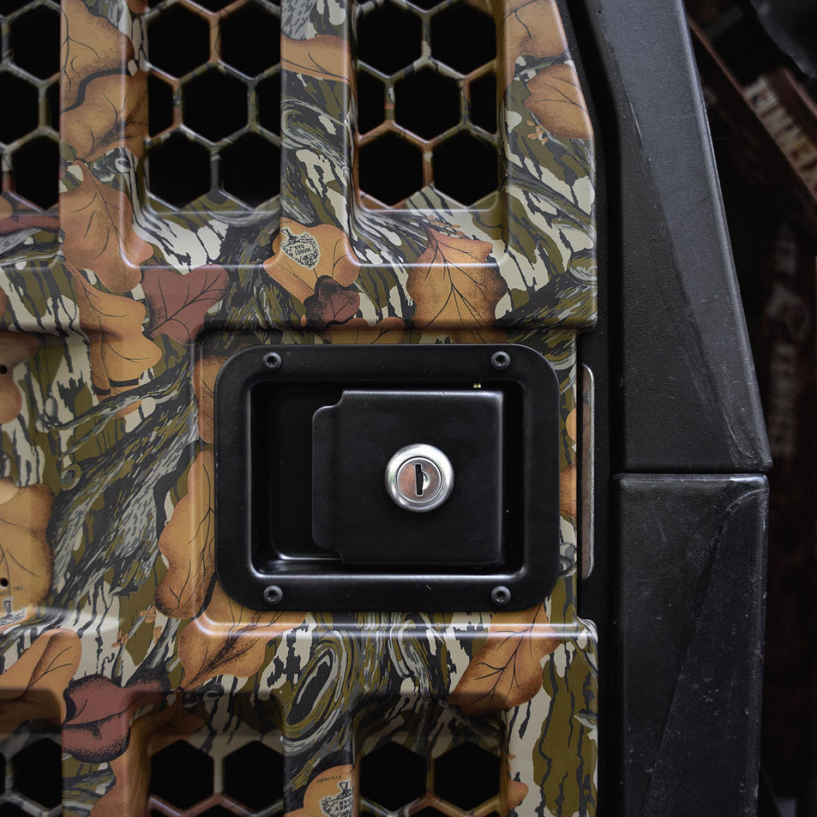 Close-up of the GUNNER G1™ KENNEL DOOR in Mossy Oak Fall Foliage camo, showing a centered black locking handle and hexagonal ventilation holes. A portion of the black frame appears on the right.