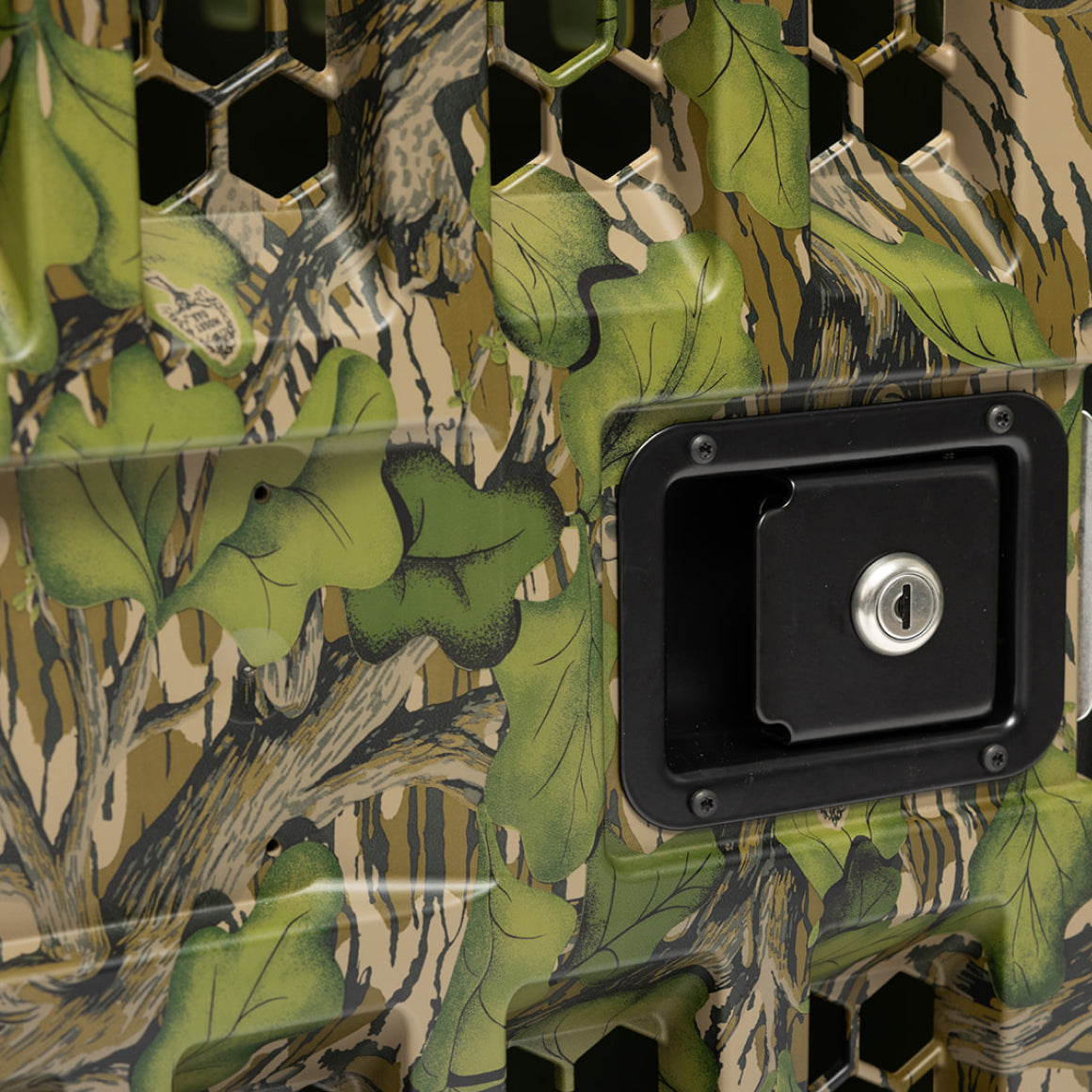 A close-up of the GUNNER G1™ KENNEL DOOR MOSSY OAK FULL FOLIAGE, featuring a leaf and tree camouflage pattern, hexagonal ventilation holes, and a rectangular black lock with a keyhole.