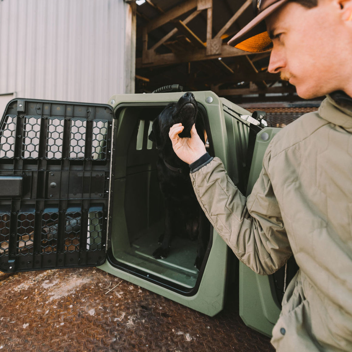 A man in a green jacket and hat gently pets a black dog sitting inside an open GUNNER G1™ KENNEL in Gunner Green, outdoors near a wooden and metal structure.