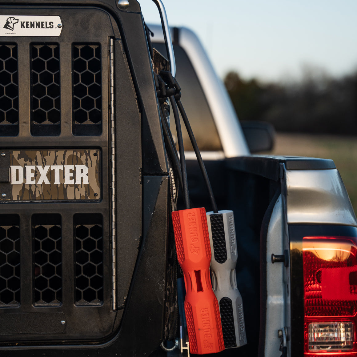 A close-up shows a GUNNER PERSONALIZED KENNEL NAME PLATE reading DEXTER on a dog crate in a truck bed, with red and gray training bumpers hanging nearby. The truck is parked outside with trees softly blurred in the background.