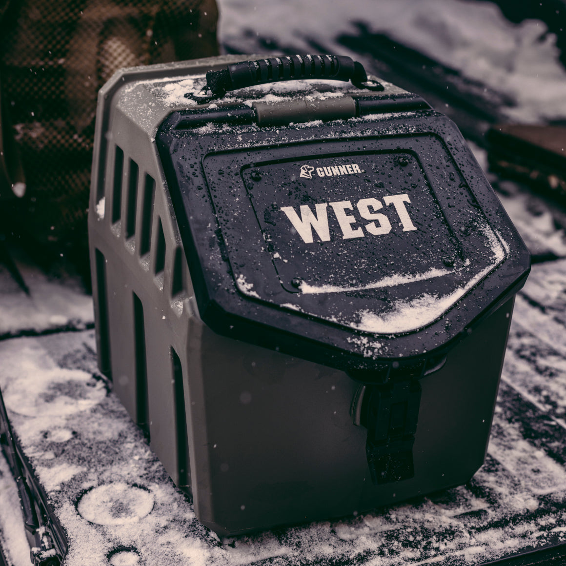A GUNNER FOOD CRATE with a handle and latch sits on snowy ground, snowflakes dusting its lid—a custom FOOD CRATE NAME PLATE adds a personalized touch to this sturdy pet carrier.