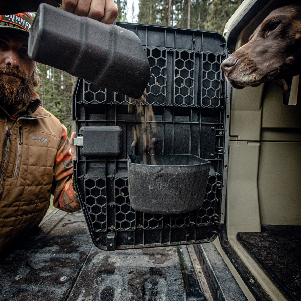 A man pours dog food into a KENNEL BOWL attached to the door of a G1 Kennel, as a brown dog watches from inside the crate outdoors near a vehicle.
