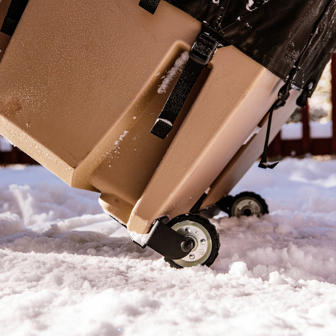 Close-up of a GUNNER KENNEL WHEEL KIT in brown, featuring black wheels and straps, rolling on snowy ground with snow sticking to the sides. A wooden fence is blurred in the background.
