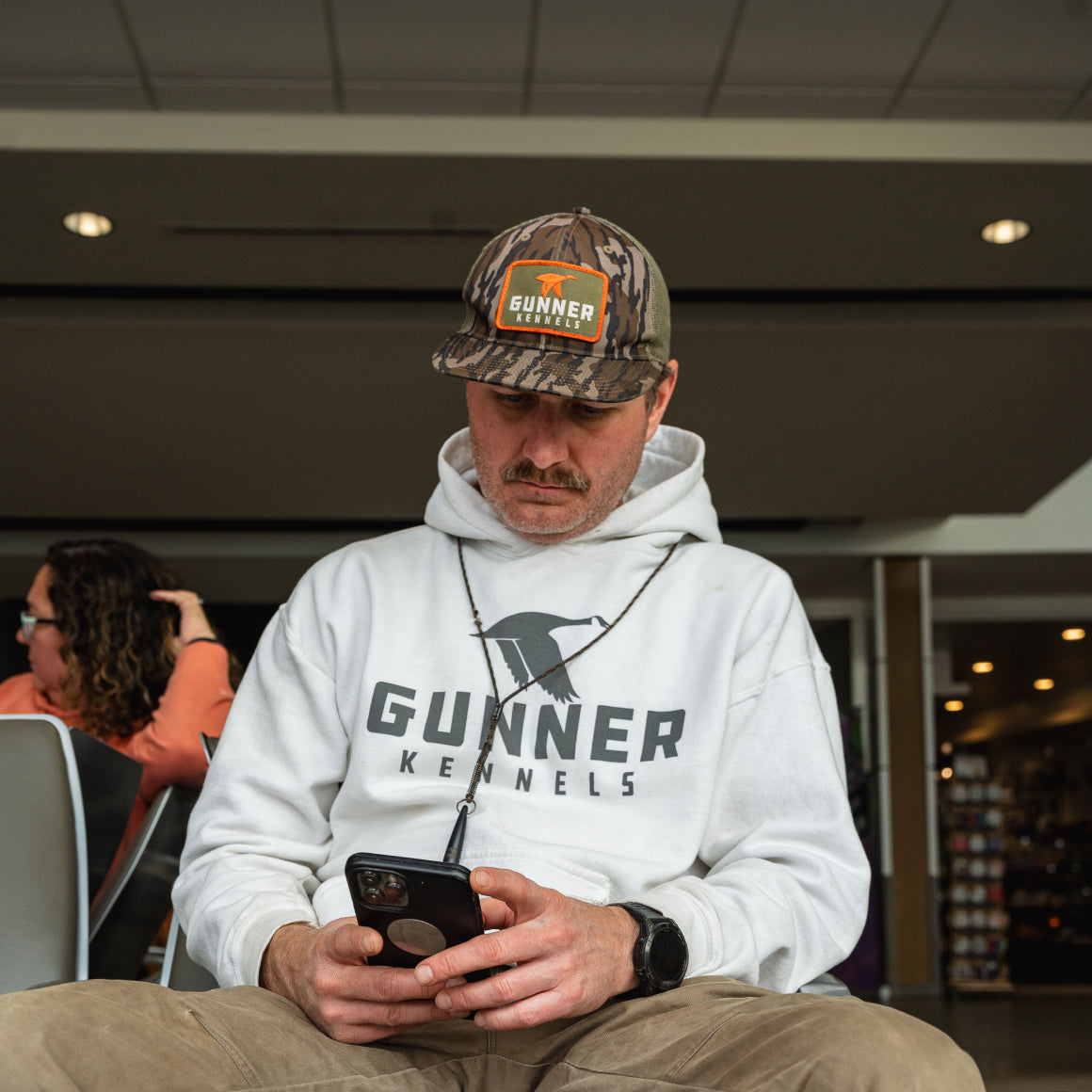 A man wearing a camouflage Gunner Kennels cap and a white GUNNER OG HOODIE sits indoors, looking down at his phone. Another person is seated in the background.