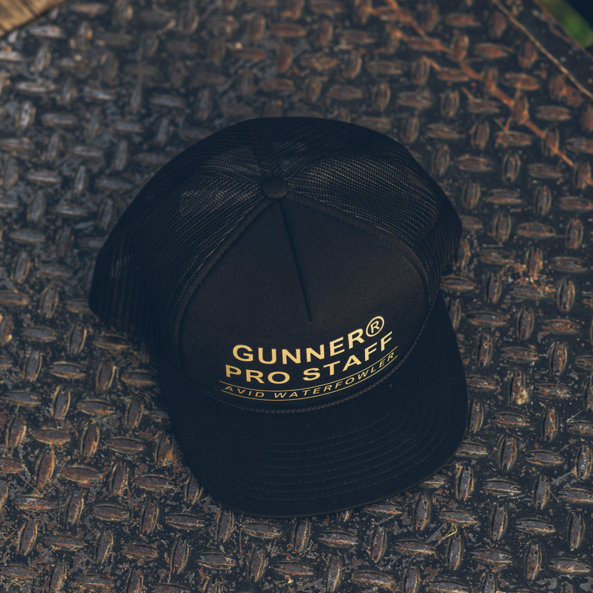 A black GUNNER PRO STAFF TRUCKER hat with mesh back and snapback closure sits on a textured metal surface, featuring yellow GUNNER® PRO STAFF AVID WATERFOWLER text on the front.