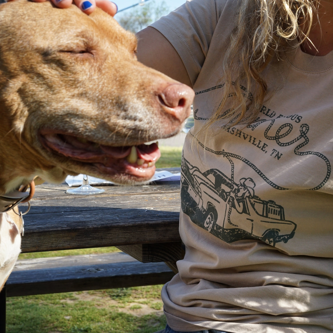 Wearing the tan WORLD FAMOUS TEE by GUNNER, a person shows genuine Southern hospitality while petting a happy brown and white dog outdoors at a wooden picnic table in the sunlight.