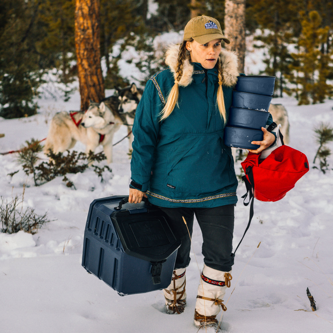 A woman in winter gear carries the GUNNER DOG BOWL - NAVY by GUNNER, along with containers and a red bag, through snowy woods. Huskies stand among trees in the background.