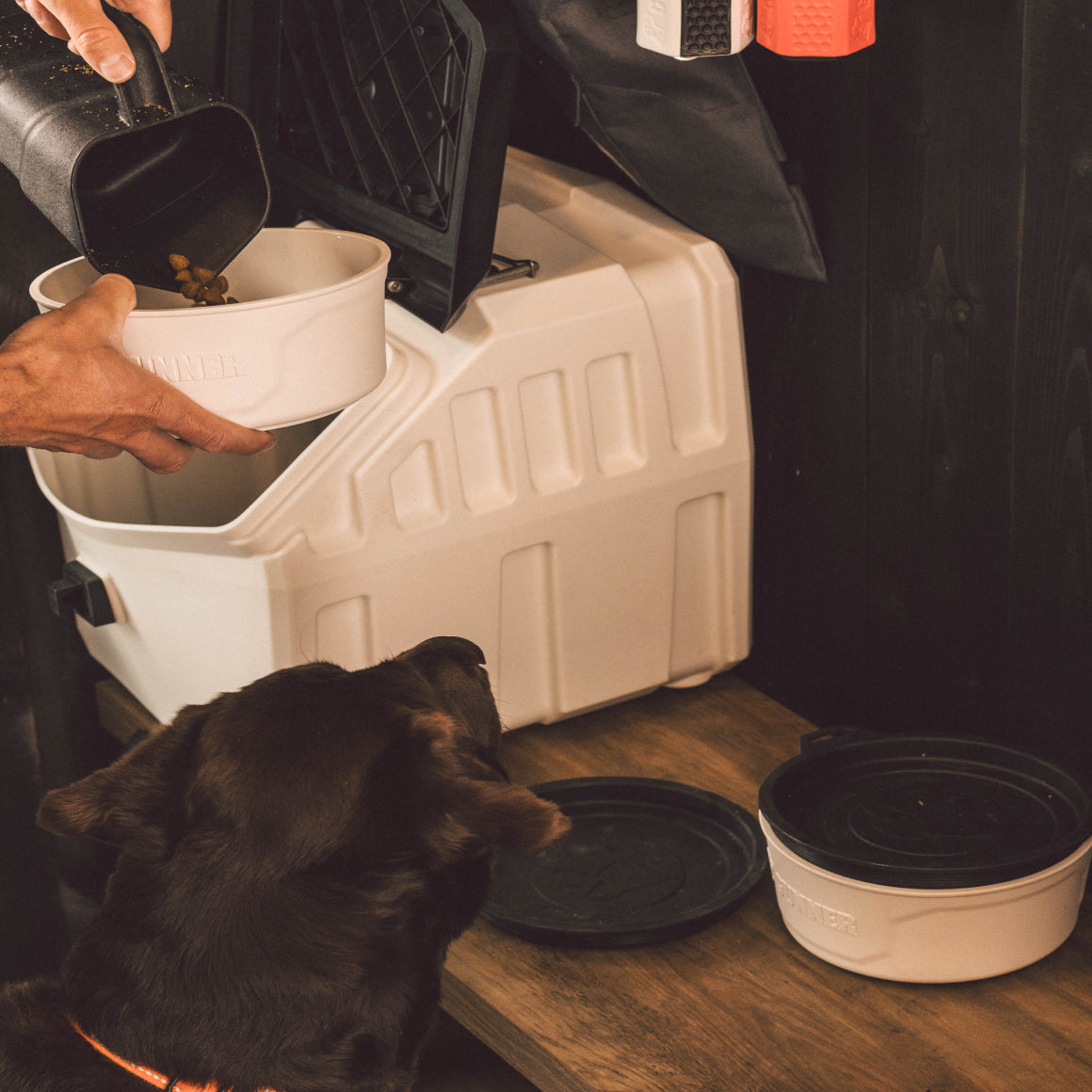 A person pours dog food from a scoop into a GUNNER DOG BOWL - GUNNER YARD by GUNNER while a brown dog watches. The bowl is in front of a white, American-made pet food storage container with black spill-proof lids nearby.
