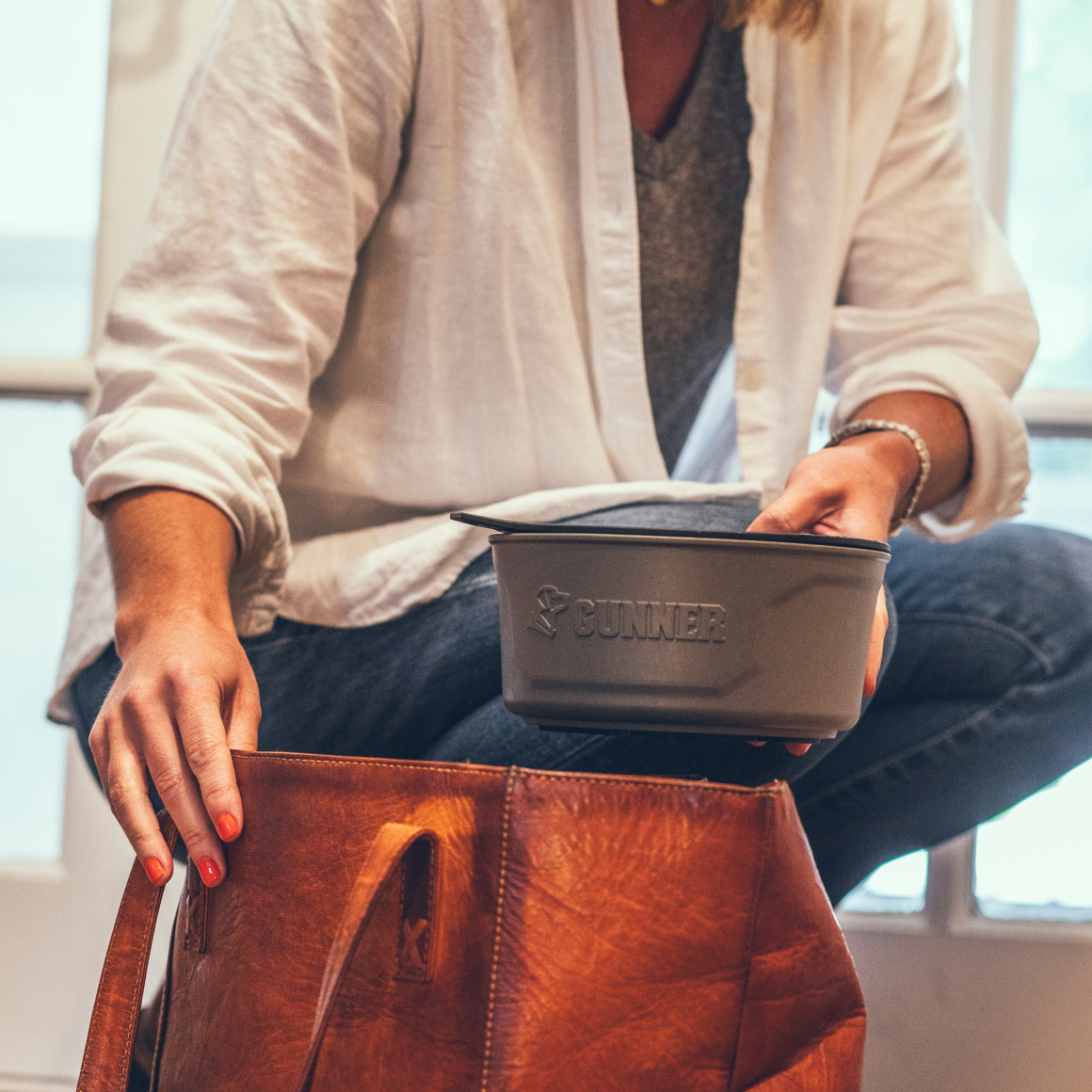 A person in a white shirt kneels indoors, placing a GUNNER DOG BOWL - GUNMETAL by GUNNER into a large brown leather bag, with sunlight streaming through the window in the background.