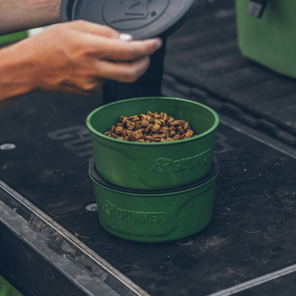 A hand lifts the lid off a GUNNER DOG BOWL - DUCK GREEN by GUNNER, filled with dry pet food, with another empty green bowl stacked beneath it. Both bowls rest on a black outdoor surface.