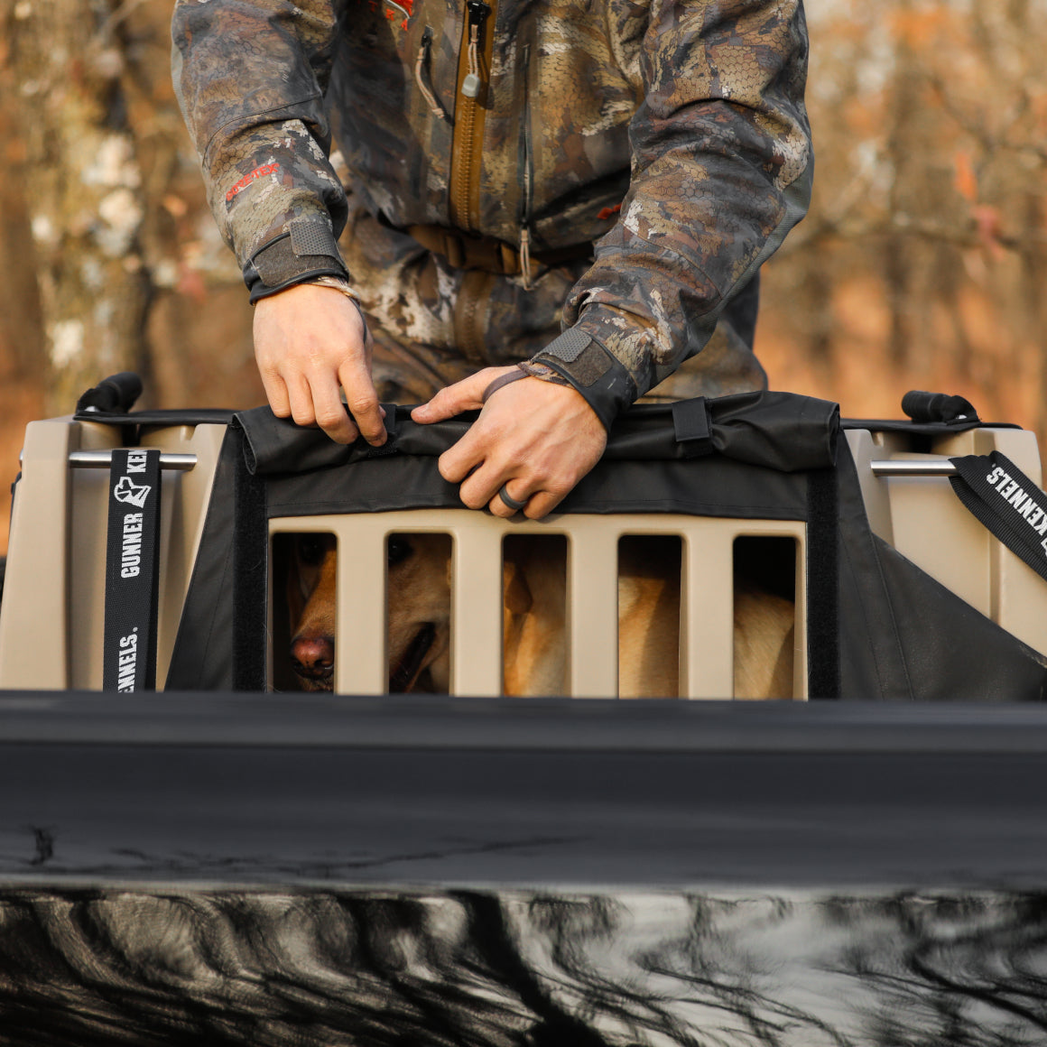A person in outdoor gear secures a cover over a tan dog crate with a dog inside, visible through the crate’s bars. The crate rests on a black surface outdoors, with blurred trees in the background.