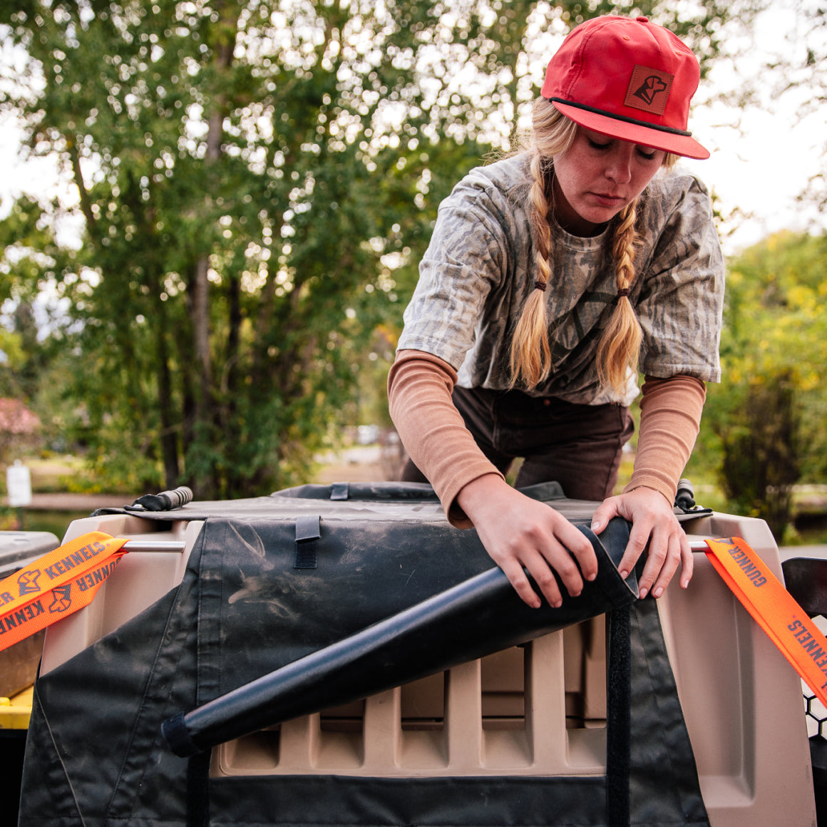 A woman in a red cap and GUNNER BOTTOMLAND TEE fastens gear to a large tan and black crate with orange safety flags, outdoors among trees in a Mossy Oak Bottomland setting.