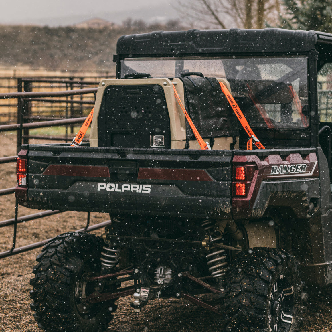 A Polaris Ranger utility vehicle parked outdoors in the rain with a large cooler and a GUNNER kennel featuring a COLD WEATHER DOOR COVER in the cargo bed, both secured with orange straps. Muddy ground and a fence are visible behind.