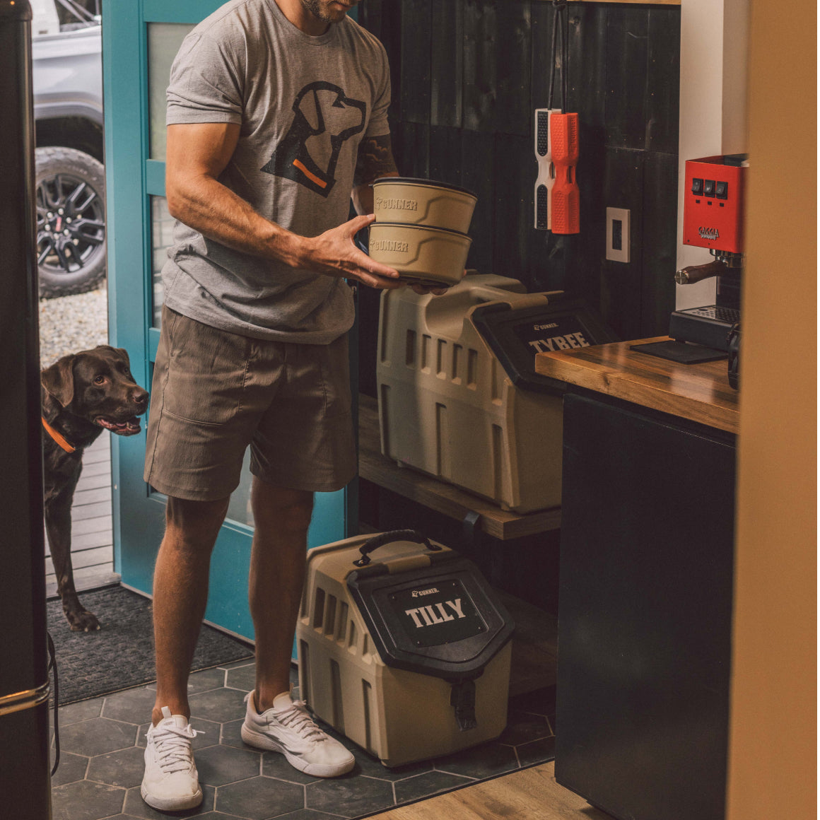 A man in a dog-themed shirt holds two labeled food containers in a kitchen, with a black dog beside him. Stacked nearby are GUNNER FOOD CRATE NAME PLATES customized with Tilly and Tyree, plus a bright red coffee machine on the counter.