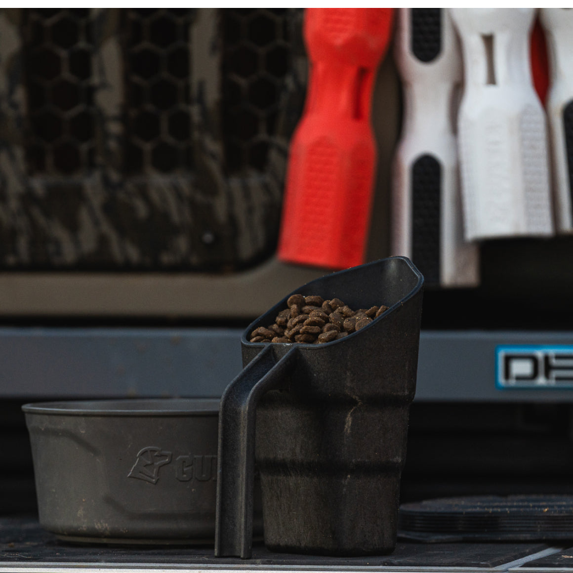 A durable GUNNER FOOD SCOOP filled with dry pet food sits in front of a gray bowl, while red and white textured dog toys hang on a rack in the background.