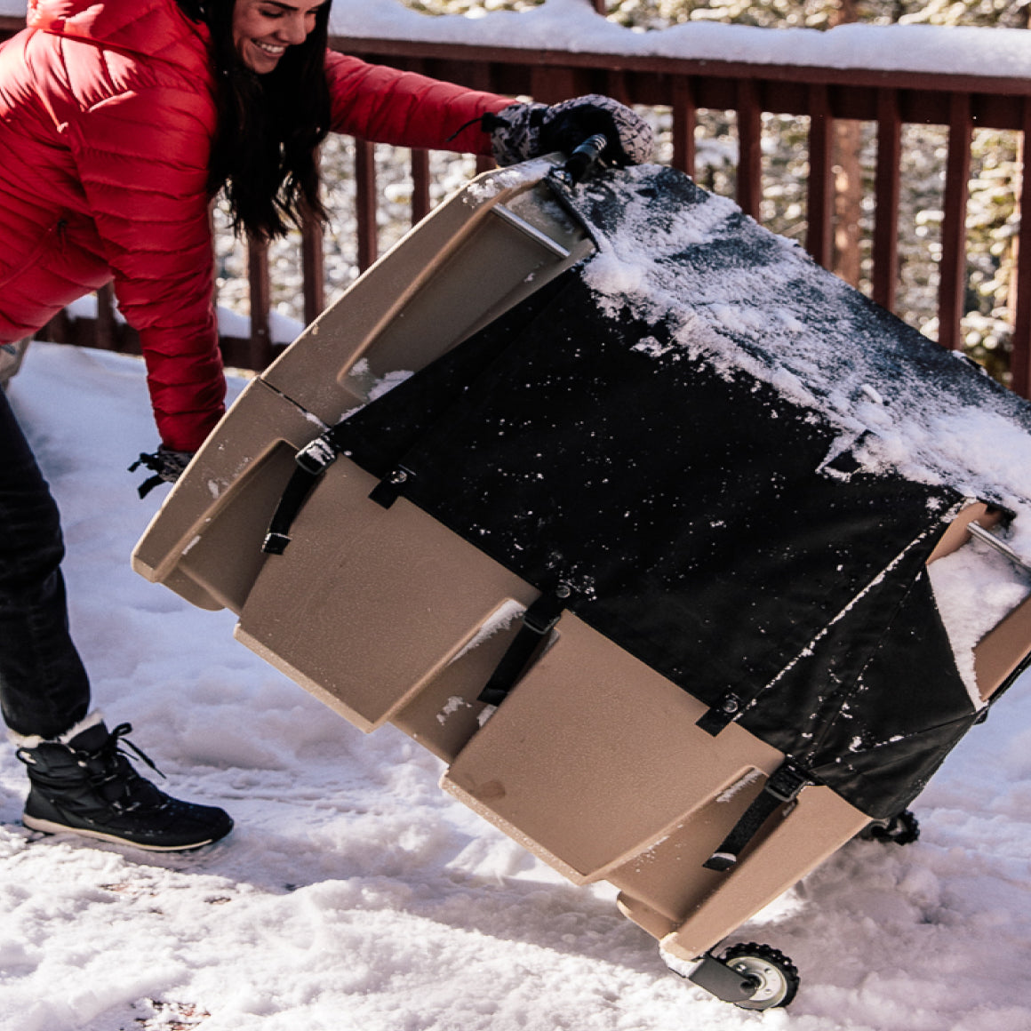 A person in a red jacket and black gloves tilts a large, wheeled brown container covered with snow outdoors on a snowy surface near a wooden railing, surrounded by outdoor gear.