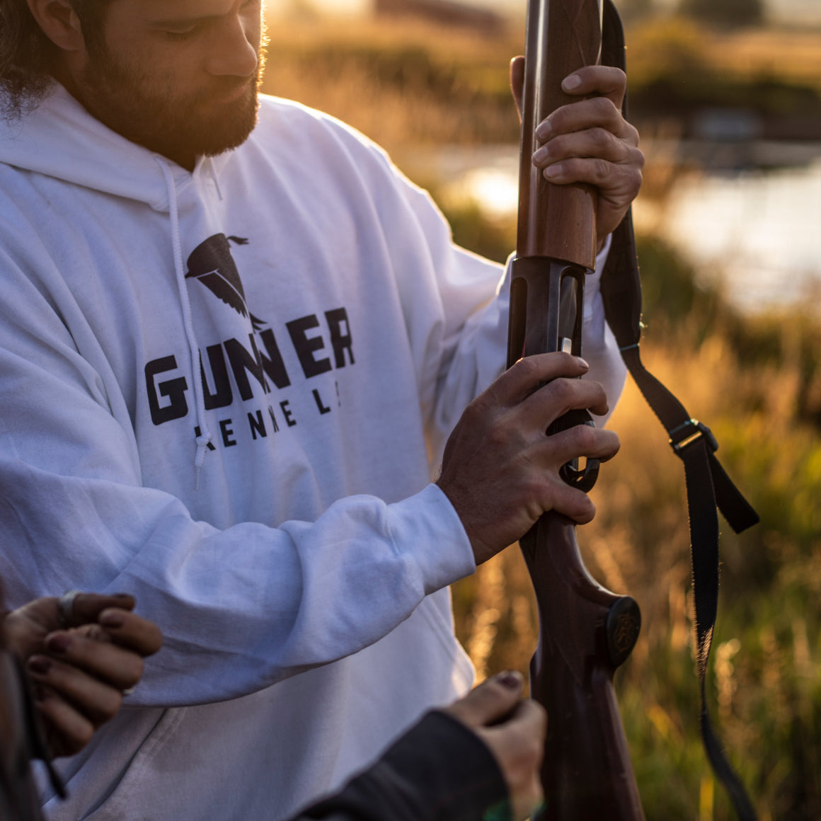 A person in a white GUNNER OG HOODIE stands outdoors by tall grass, loading a shotgun as sunlight streams in. Another individuals hands are visible in the foreground.