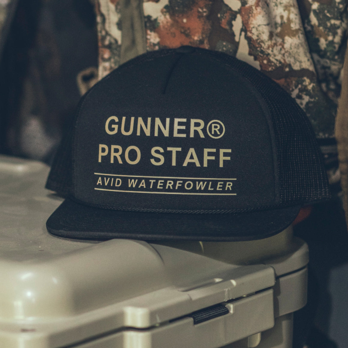 A black GUNNER PRO STAFF TRUCKER hat with bold white lettering rests on a closed light-colored plastic container, with a camo jacket in the background.