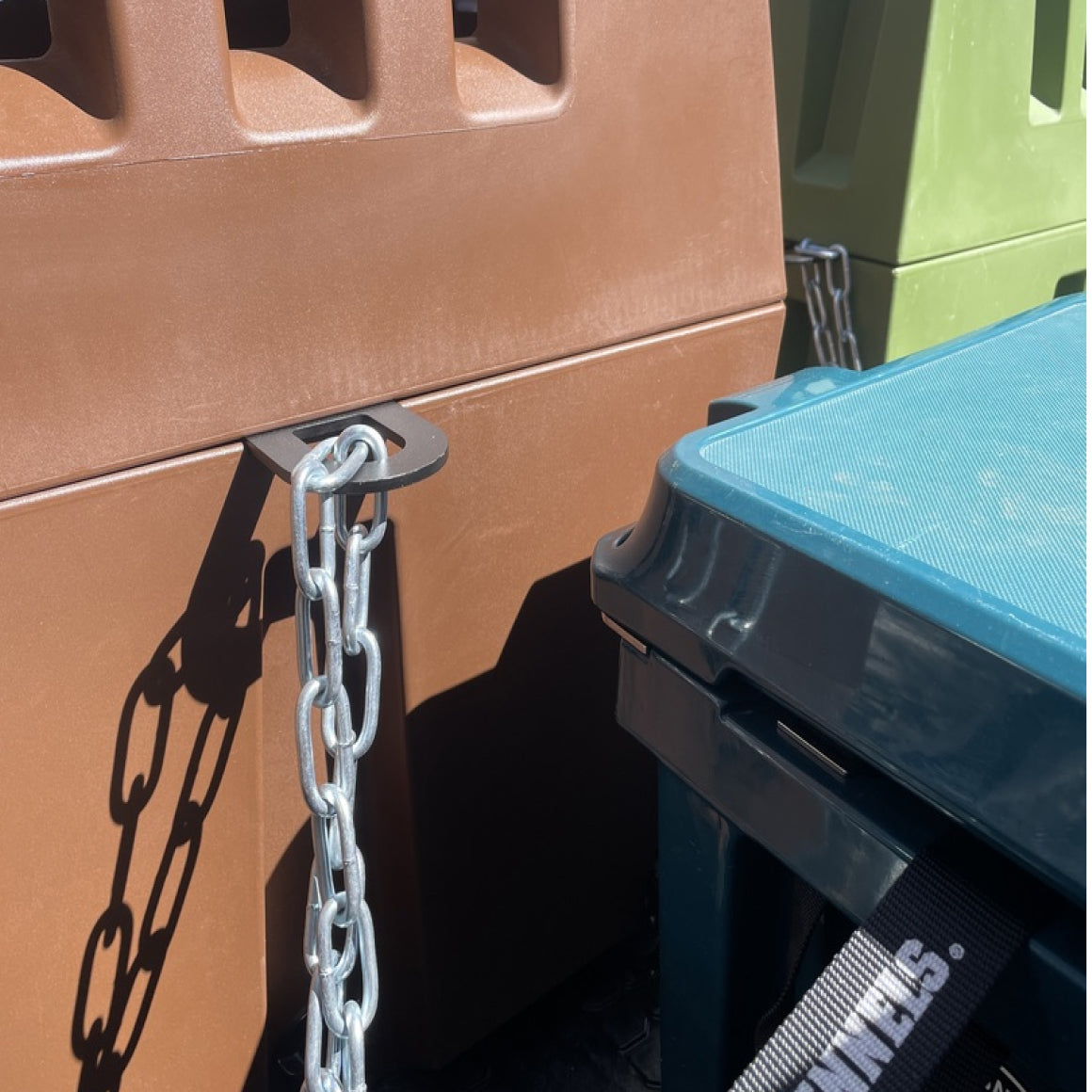Close-up of a brown trash bin, green recycling bin, and blue container—all secured with metal chains using the SECURITY LOCK KIT. Shadows and chain links stand out against the plastic surfaces in bright sunlight.