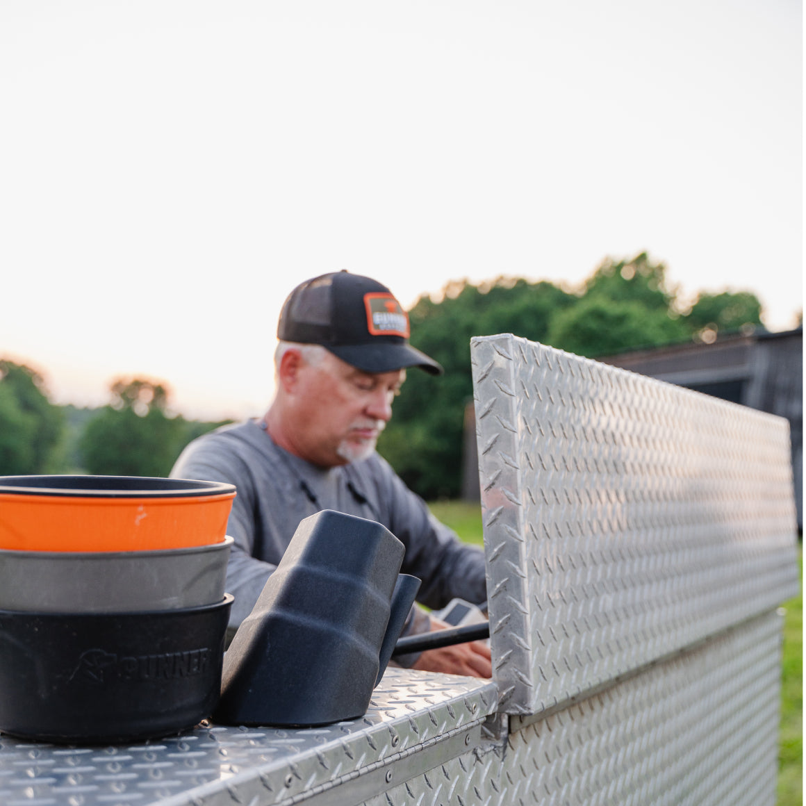 A man in a cap stands by a metal truck storage box at sunset, with GUNNER DOG BOWL - BLACK by GUNNER and a plastic scoop on the edge. Trees and grass are visible in the background.