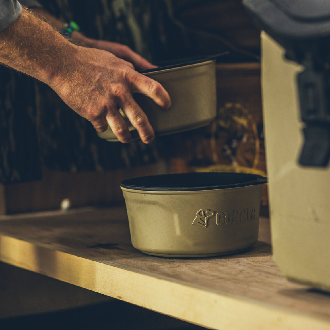 A hand sets a GUNNER DOG BOWL - TAN on a wooden surface beside another identical bowl, with the GUNNER logo clearly visible on one.