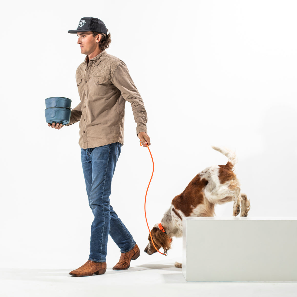 A man in a tan shirt, blue jeans, boots, and a cap walks a brown and white dog on an orange leash while holding the GUNNER DOG BOWL - Ducks Unlimited Blue near a Ducks Unlimited waterfowl habitat.