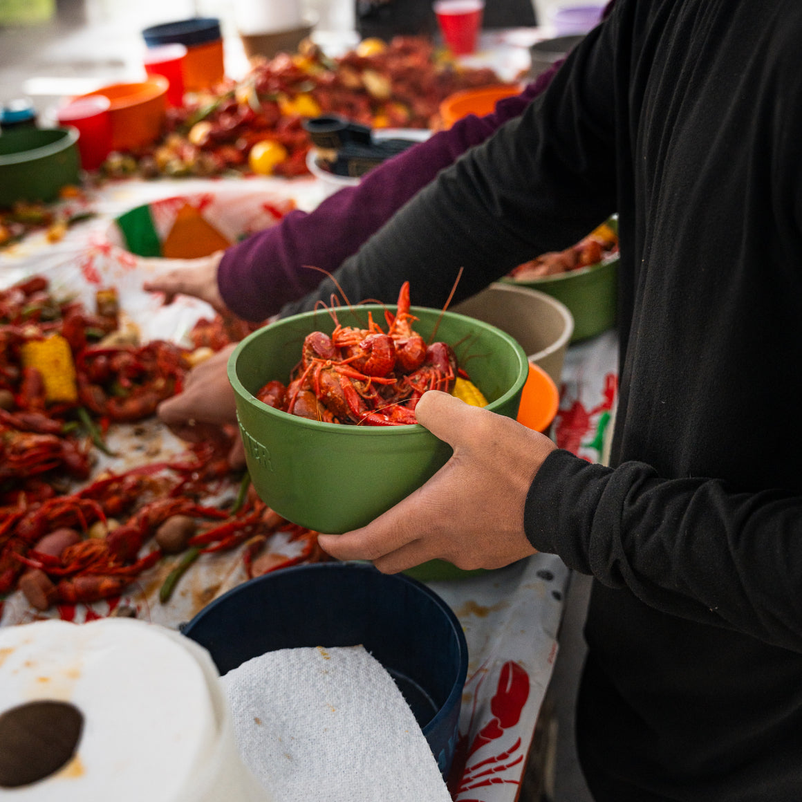 Two people serve themselves crawfish from a messy table with corn and sausage. One holds a GUNNER DOG BOWL - DUCK GREEN by GUNNER filled with crawfish, while the other reaches for food. Bowls and paper towels are nearby.