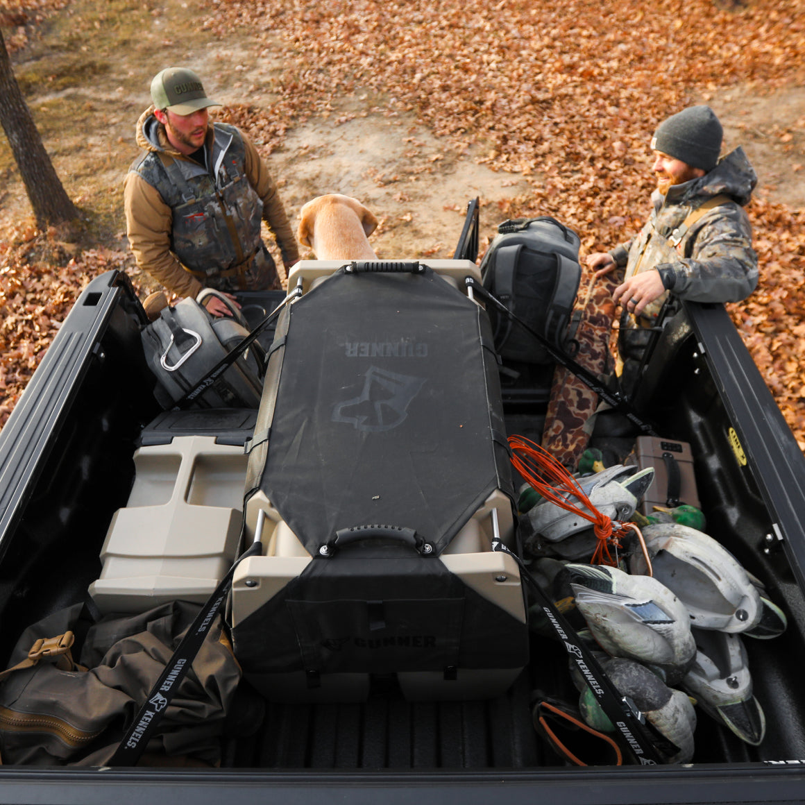Two people in outdoor gear load hunting or camping equipment and a large dog crate fitted with the GUNNER ALL-WEATHER KIT 2.0 BLACK into a pickup truck bed surrounded by autumn leaves. Various bags, decoys, ropes, and other gear—kept safe and shielded with added UV protection by the GUNNER ALL-WEATHER KIT 2.0 BLACK—are visible in the truck bed.