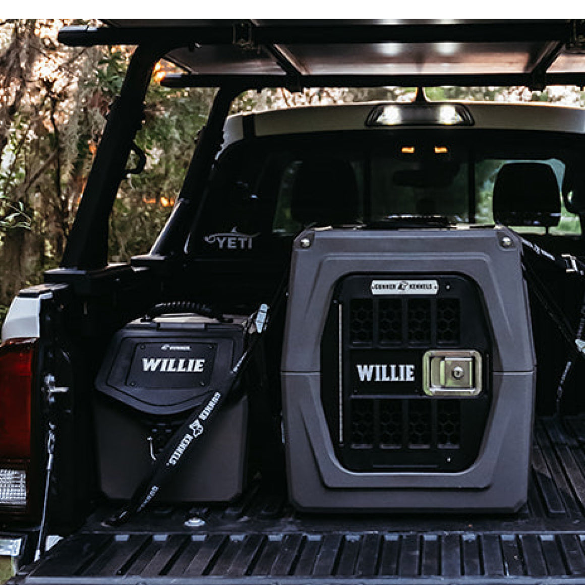 Two gray dog kennels labeled “WILLIE” are secured in the bed of a pickup truck outdoors, with the truck’s back open and trees visible in the background—ready to transport a working dog or hunting dog.