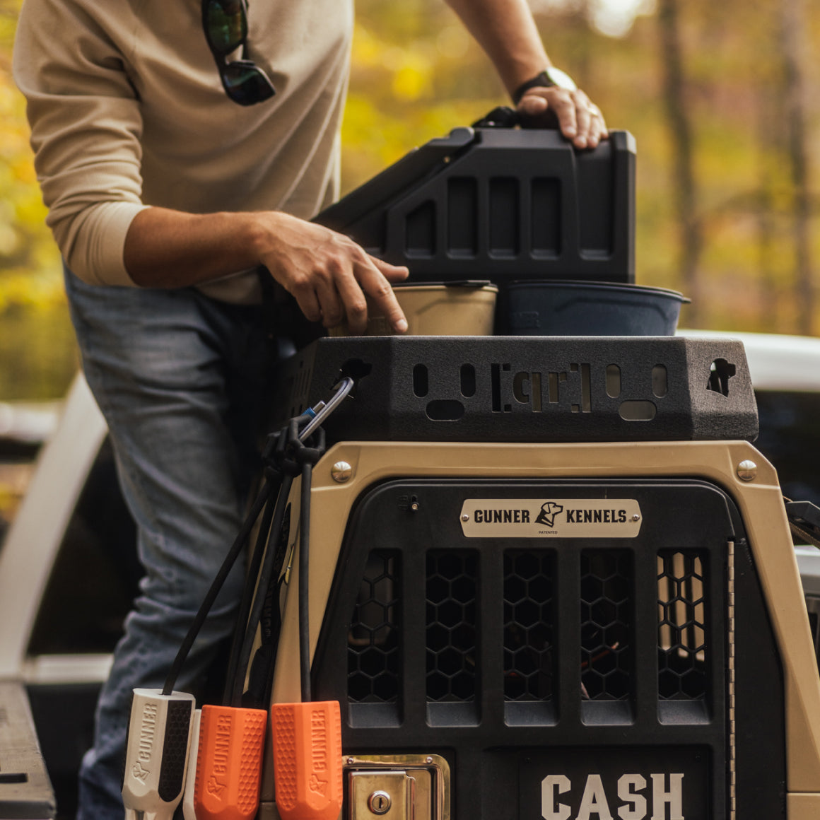 A person places a container into a pet crate labeled GUNNER Kennels and Cash in the back of a vehicle, where a GUNNER Kennel Rack BLACK by GUNNER is mounted in the truck bed as part of an organized kennel storage system. Three leash handles hang on the crate, while trees with autumn leaves appear blurred in the background.