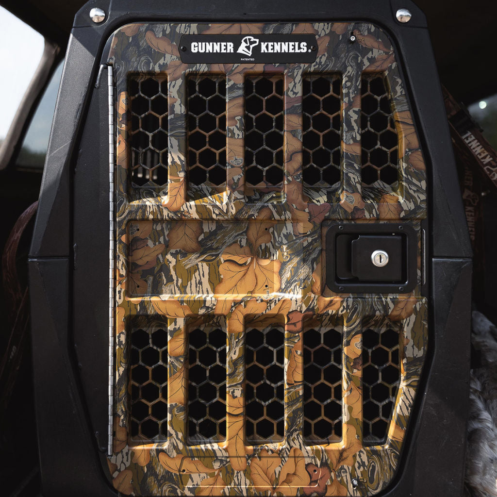 A close-up of the GUNNER G1™ Kennel Door in Mossy Oak Fall Foliage, featuring black hexagonal ventilation holes and a secure lock, shown inside a vehicle.