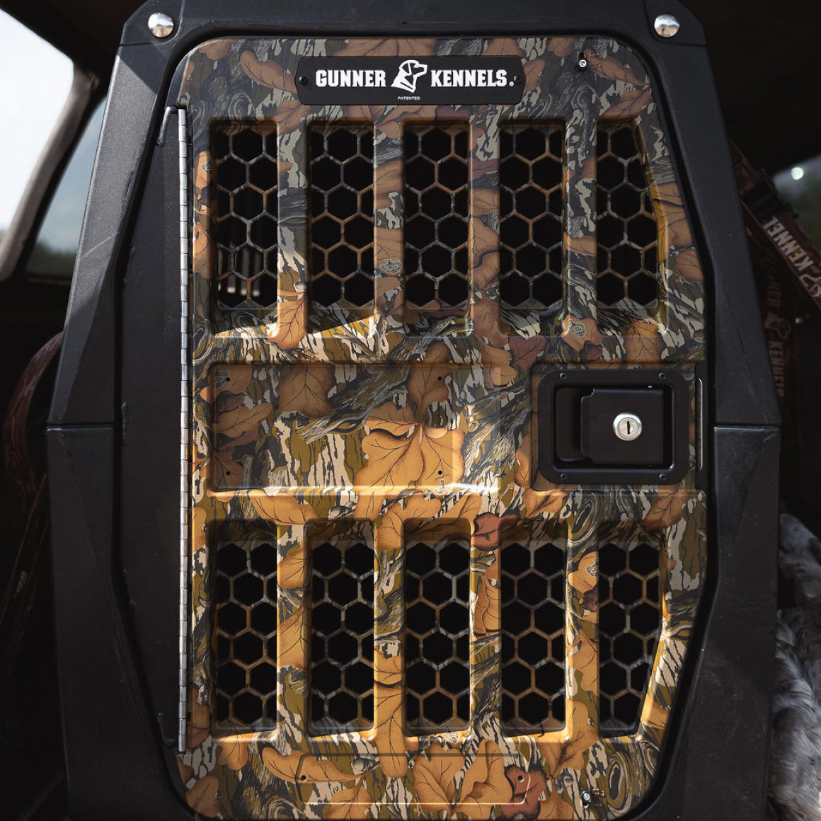 A close-up of the GUNNER G1™ Kennel Door in Mossy Oak Fall Foliage, featuring black hexagonal ventilation holes and a secure lock, shown inside a vehicle.