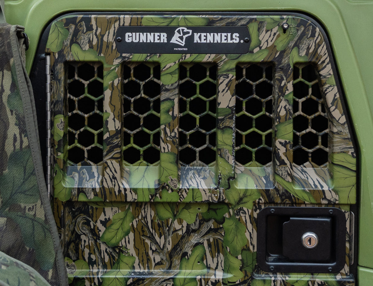 The GUNNER G1™ Kennel Door Mossy Oak Full Foliage features a camouflage pattern, hexagonal ventilation holes, a top Gunner Kennels label, and a black lock at the bottom right.
