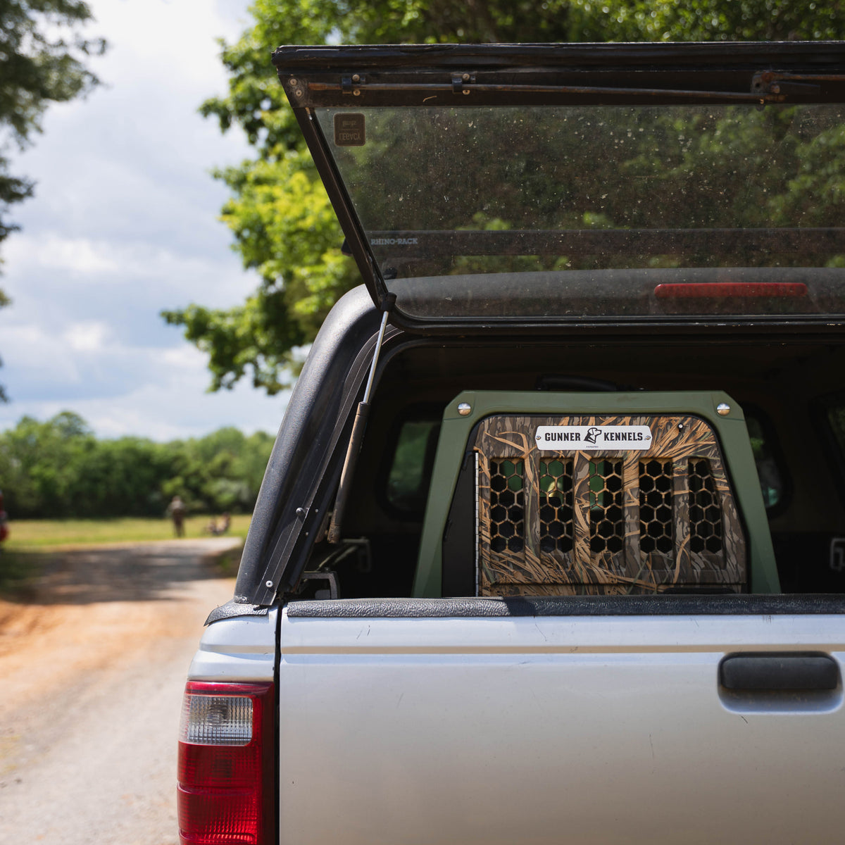 A pickup truck with its tailgate open reveals a GUNNER G1™ Kennel in Gunner Green/Mossy Oak Shadow Grass inside the truck bed, parked on a dirt road lined with trees under a partly cloudy sky.