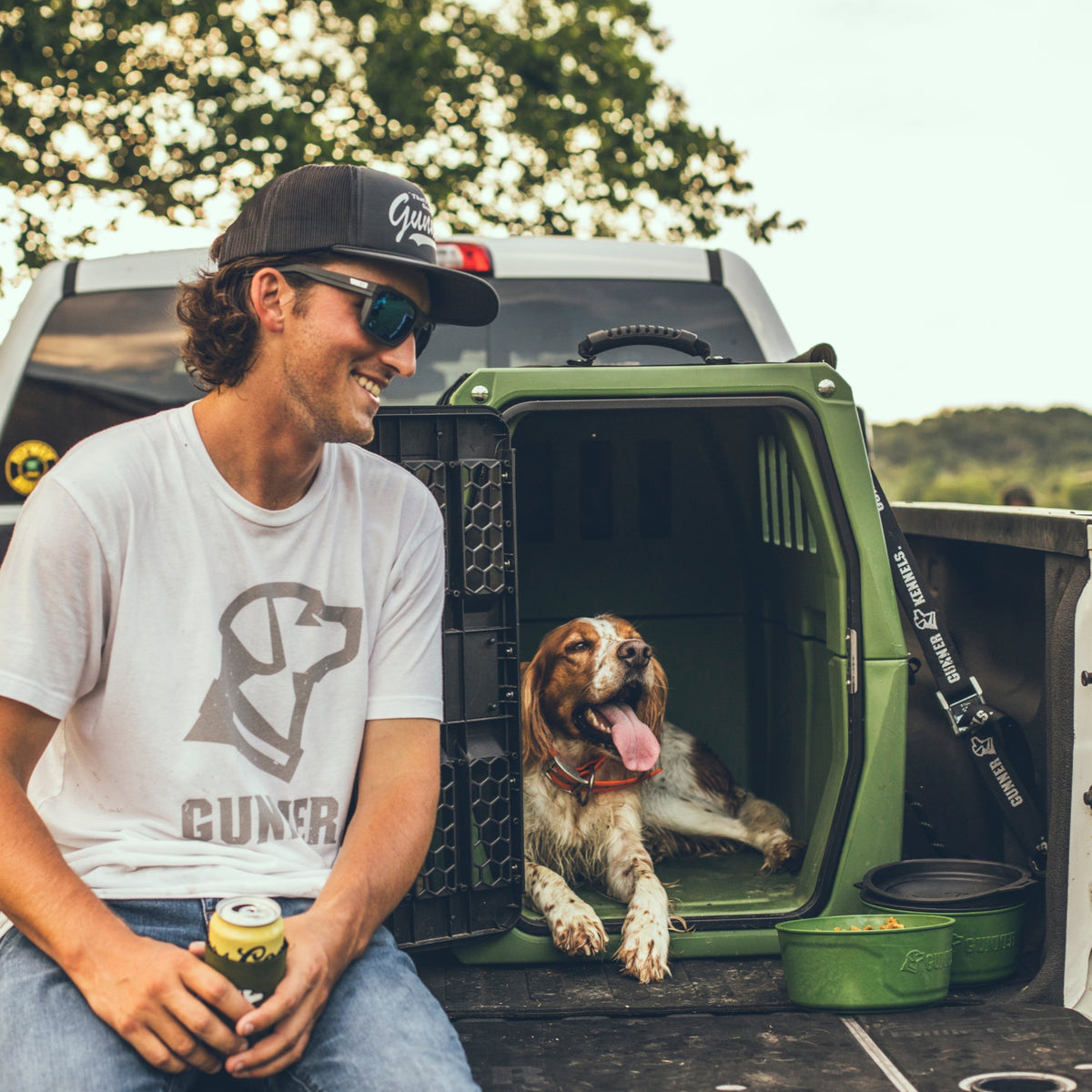 A man in sunglasses and a cap smiles on a truck bed beside a panting dog relaxing in a green crate, with the GUNNER DOG BOWL - DUCK GREEN by GUNNER nearby. Outdoor scene with trees in the background.