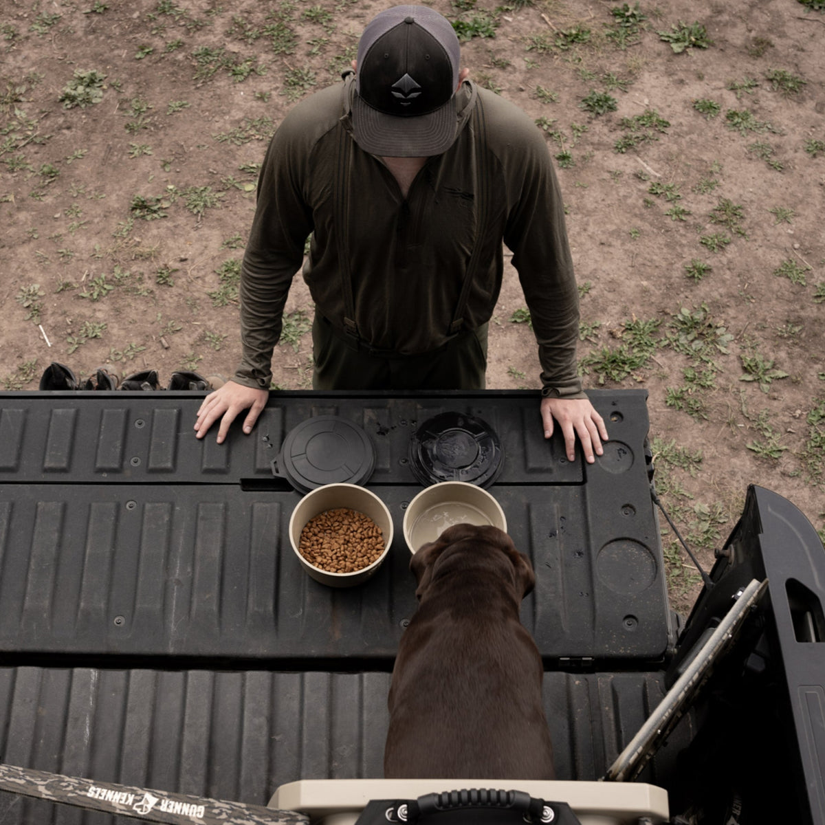 A man stands by his truck’s open tailgate watching his dog eat from a GUNNER DOG BOWL - TAN by GUNNER, with another bowl and a drink nearby on the grass and dirt outdoors.