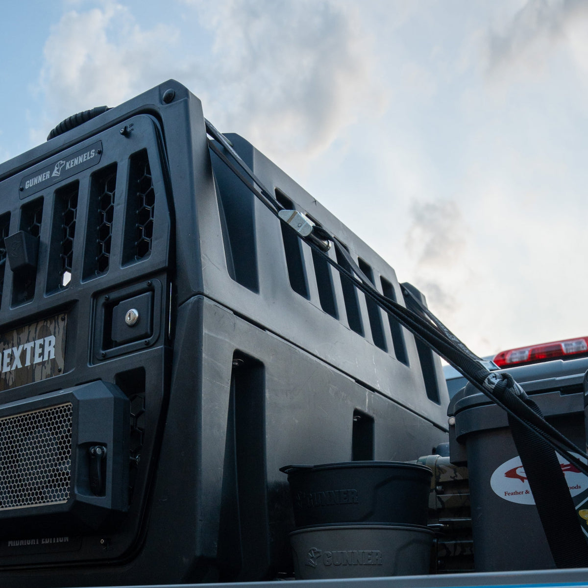 A sturdy black dog crate is strapped in a truck bed beside a gray bucket, plastic containers, and the GUNNER DOG BOWL - BLACK by GUNNER, all under a cloudy sky.