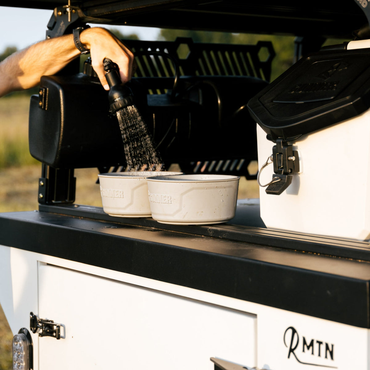 A person fills two white, American-made GUNNER DOG BOWLs by GUNNER at a black and white outdoor kitchen attached to a vehicle. Nearby are a black container, a white cooler, and trees in the background.