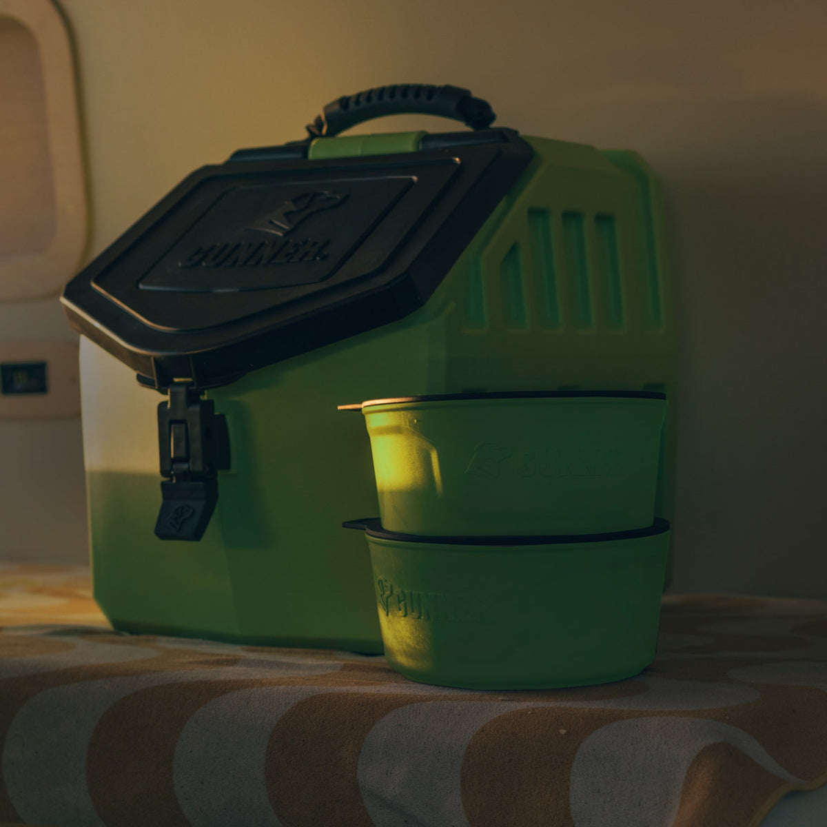 A limited-edition GUNNER Dog Bowl in Sulphur Springs and a matching green cooler with a black lid, both branded “GUNNER,” are arranged on a beige and white patterned tablecloth under warm indoor lighting.